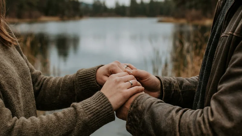 Two people walking hand in hand at the edge of still water, jackets on against an overcast sky, the quiet between them saying as much as any words