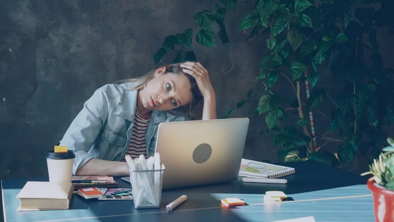Person at a wooden desk in early morning light, staring at a blank laptop screen with an untouched coffee beside them, expression of foggy frustration