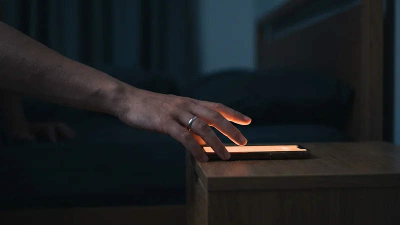 A hand reaching for a phone on a nightstand in a darkened bedroom, the screen the only source of light in the room.