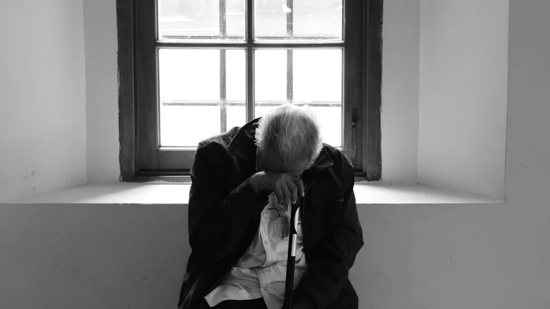 An elderly person and a young adult sitting across from each other at a kitchen table, afternoon light, both holding mugs