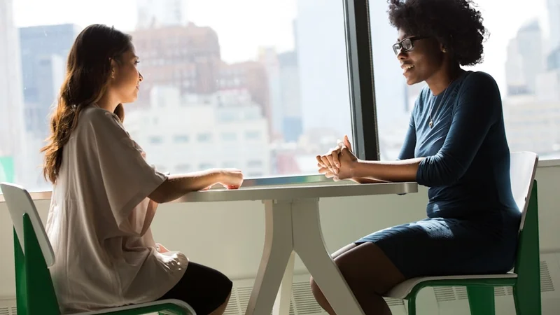 Two people in a candid, honest conversation — one leaning in, both visibly relieved, the impostor experience losing its power through connection and shared recognition