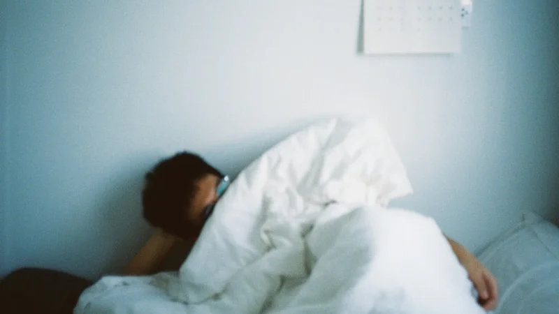 Person sitting on the edge of a bed in morning light, rubbing their eyes, alarm clock showing 8:00 behind them — expressing exhaustion despite a full night's sleep