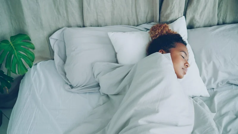 Minimalist bedroom optimised for sleep — blackout curtains drawn, warm dim light, sunrise alarm clock and brown noise machine on nightstand